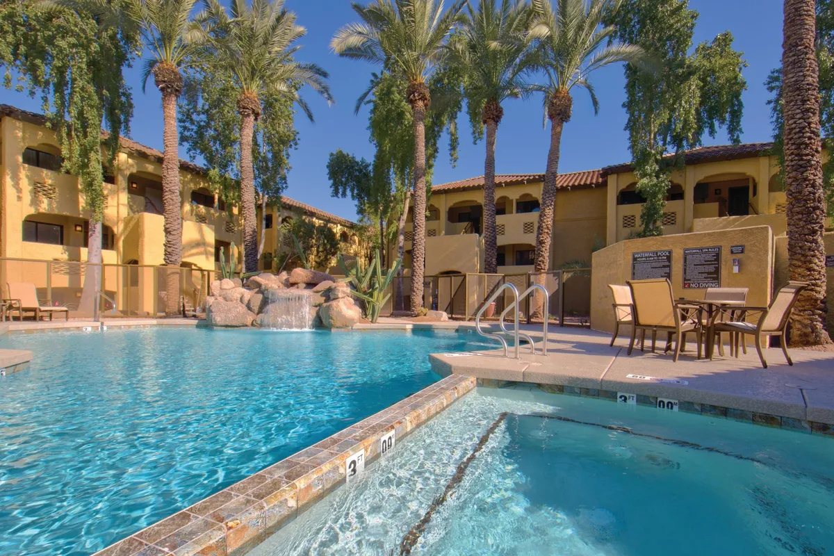 This image showcases a tranquil and luxurious swimming pool area surrounded by palm trees and patio seating. Waterfall features add a sense of movement and relaxation, while people are seen swimming, sunbathing, and socializing by the poolside. The sunset backdrop adds warmth and atmosphere, elevating the resort's inviting ambiance.