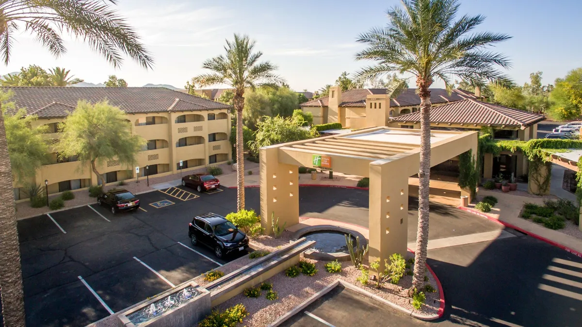 The image showcases the exterior of the property, framed by tall palm trees and an inviting entrance canopy. A tranquil water feature adds a touch of relaxation, while guests can be seen arriving and gathering outdoors, suggesting a welcoming and social environment. The mood created is both inviting and serene, ideal for relaxation after a day of exploring or golfing.