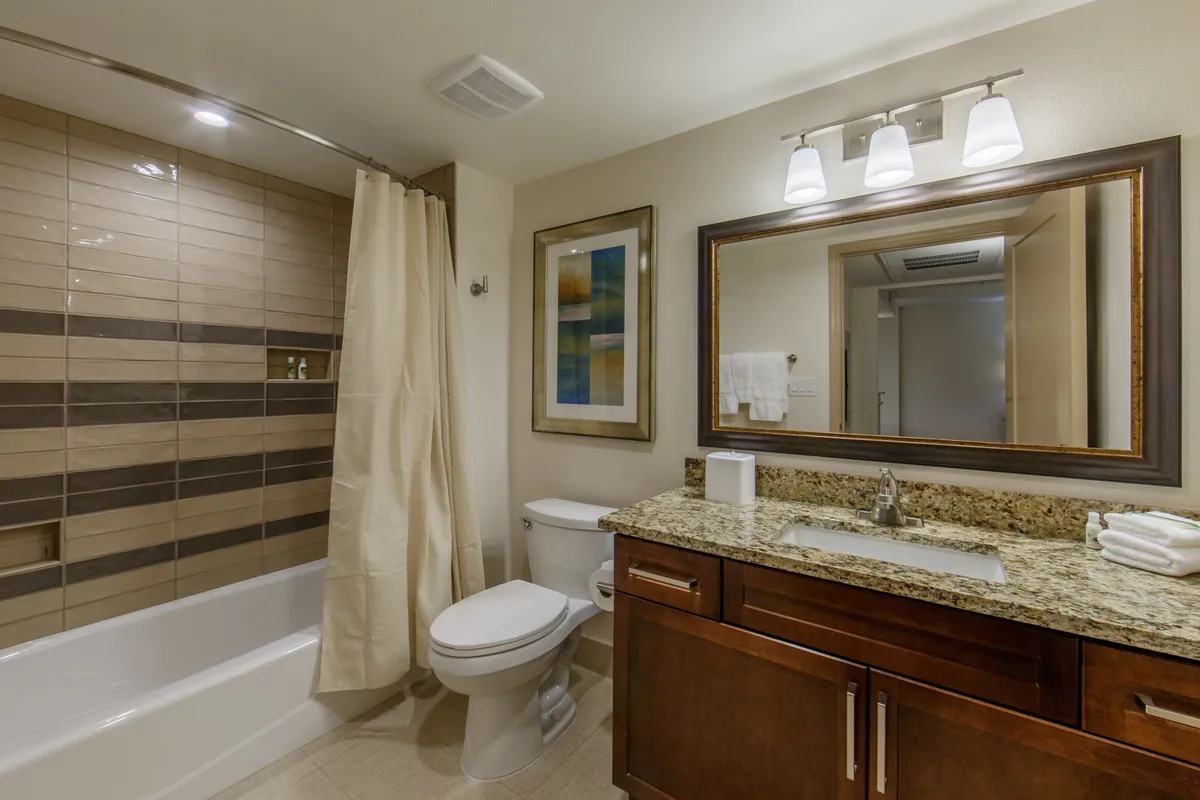 The image presents a modern bathroom with a granite countertop, vanity mirror, inviting shower area, and tasteful framed artwork. The space appears fresh and well-equipped for unwinding or getting ready, with ample towels and toiletries displayed.