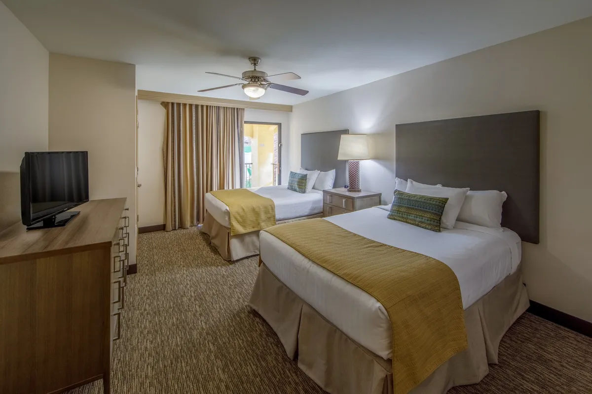 A bright bedroom with two queen-sized beds, a nightstand with a lamp, a television, and a window with curtains. The scene is comfortable and welcoming, showing a well-appointed area for relaxation and unwinding, which hints at the privacy and personal comfort available in the villa.