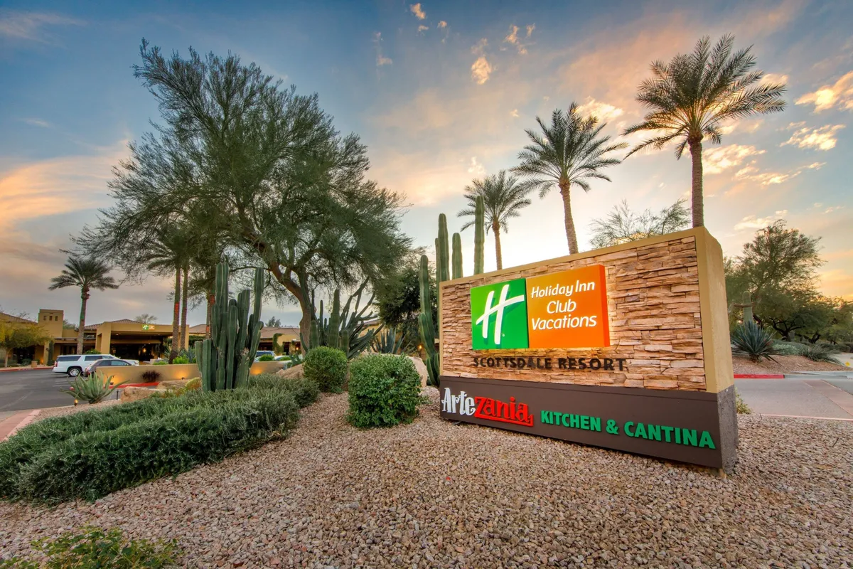 An inviting exterior shot of the resort showcases the iconic Holiday Inn sign, surrounded by desert landscaping and tall palm trees. The image hints at the resort's proximity to area attractions and highlights the welcoming atmosphere for guests preparing to explore Scottsdale's treasures.