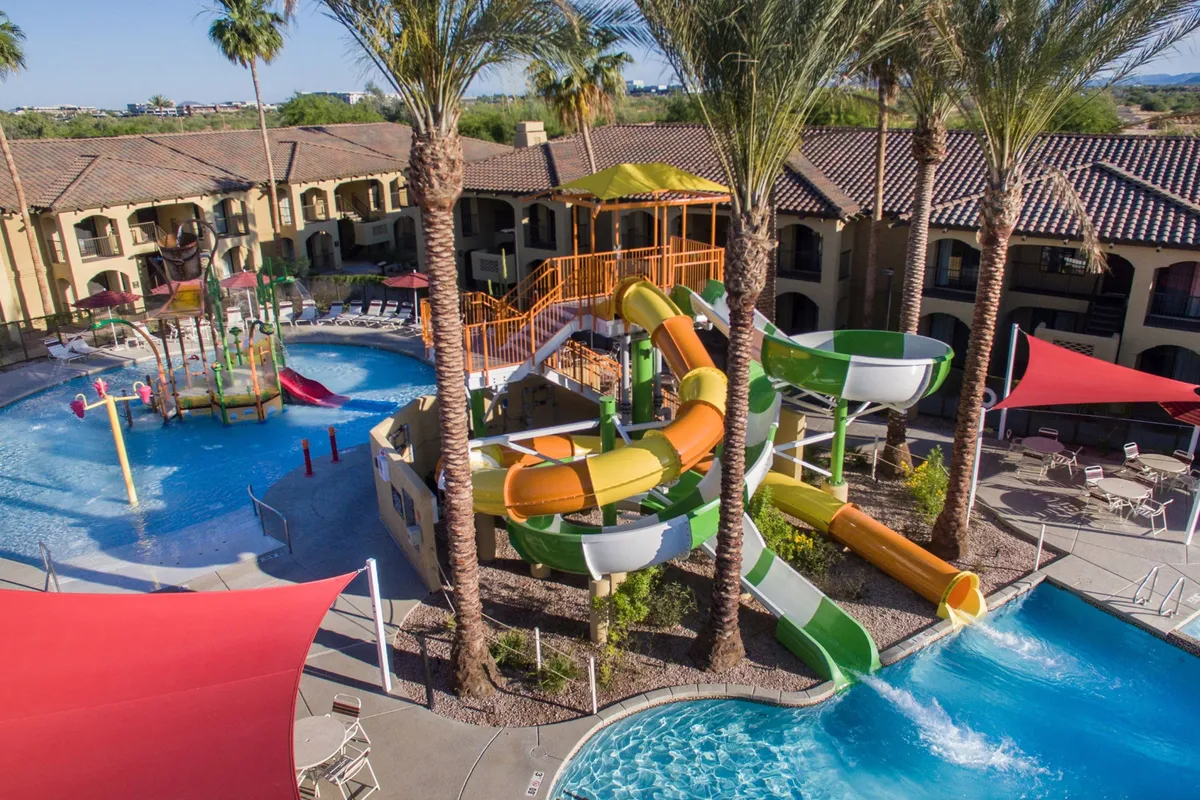 This vibrant exterior scene captures the resort's swimming pool area with exciting water slides, happy families, lush palm trees, and a playful atmosphere. Children are splashing in the heated pool while others are ready to zoom down the slides, perfectly embodying the spirit of family fun and outdoor adventure.