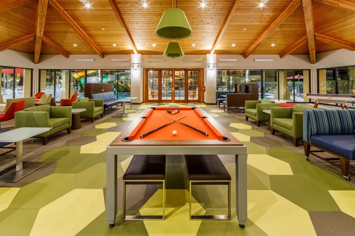 This interior shot features a stylish room with a pool table, hexagonal patterned flooring, green armchairs, large windows, and a wooden ceiling. The space exudes a playful yet relaxed atmosphere ideal for unwinding or spending time with friends and family.