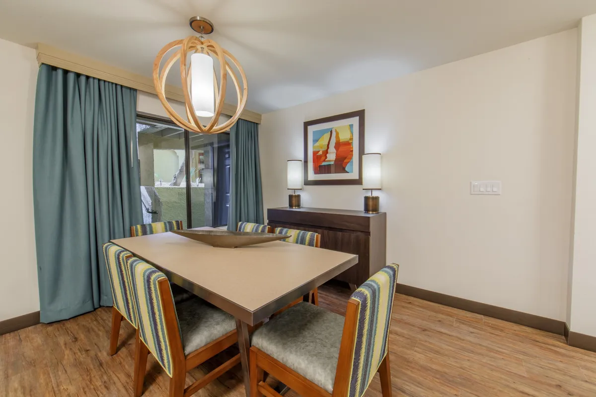 A modern dining area with a large table, decorative pendant light, colorful artwork, a window with curtains providing natural light, and a sideboard with lamps. The setup is perfect for shared meals, casual gatherings, or even game nights, emphasizing group interaction and the villa's social nature.